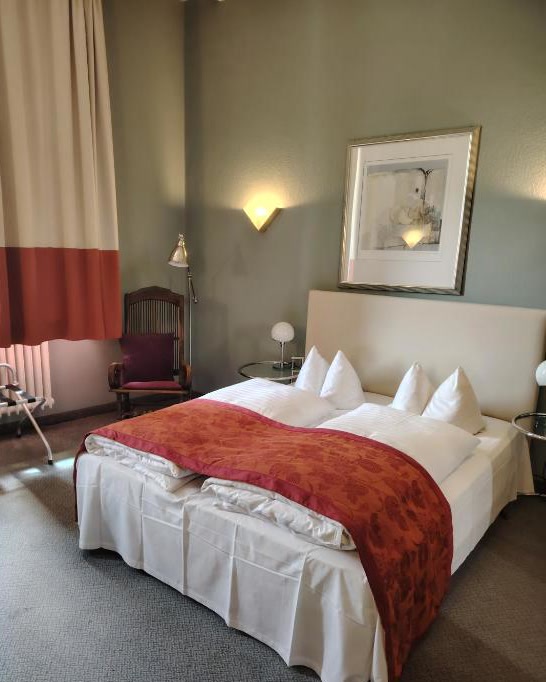 Doppelzimmer mit salbeigrünen Wänden, Polsterbett und terrakottafarbenem Bettläufer vor hohem Fenster